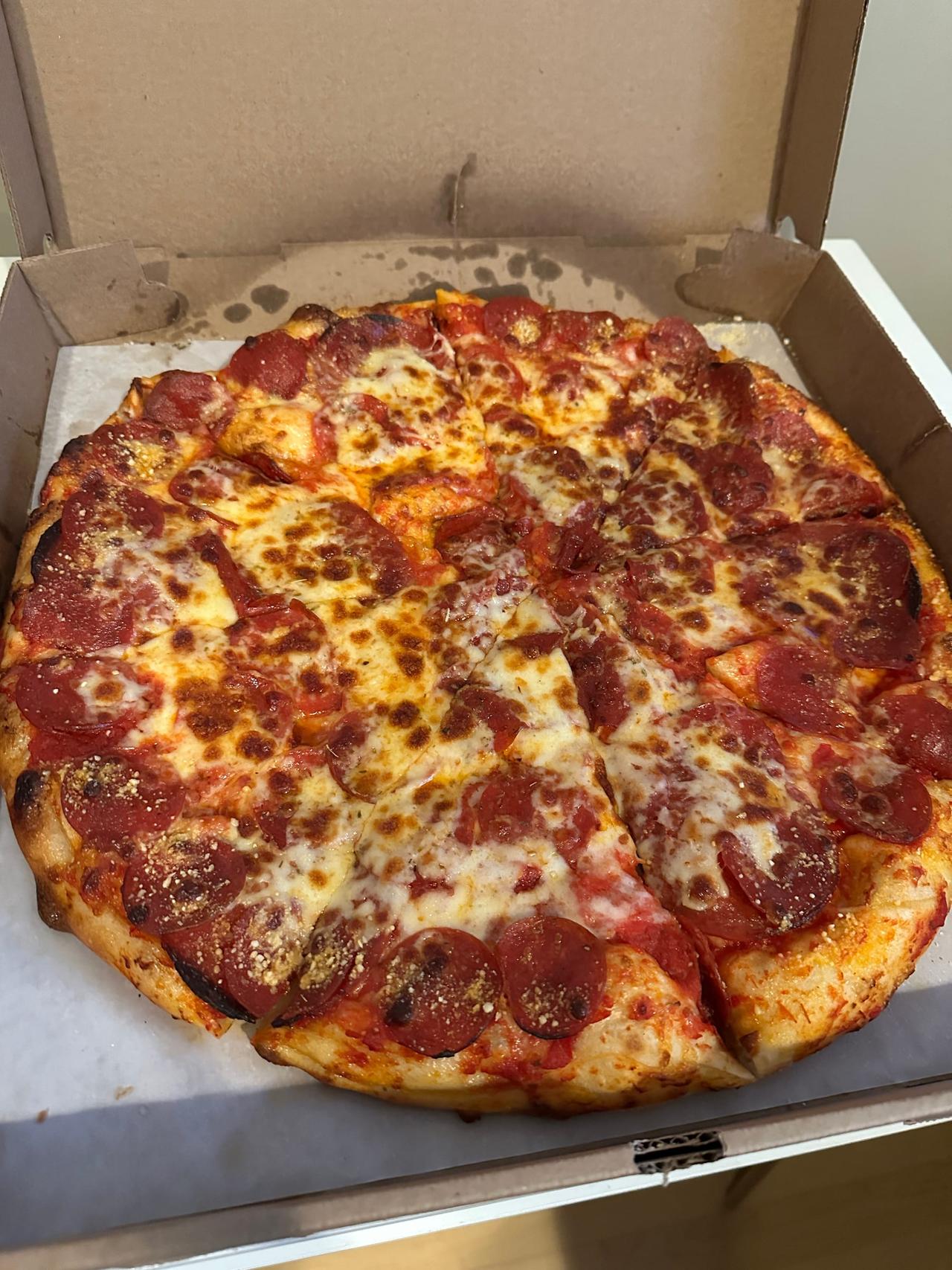 Pepperoni pizza with browned, bubbly cheese and a seasoned crust edge, sitting in a plain cardboard box.